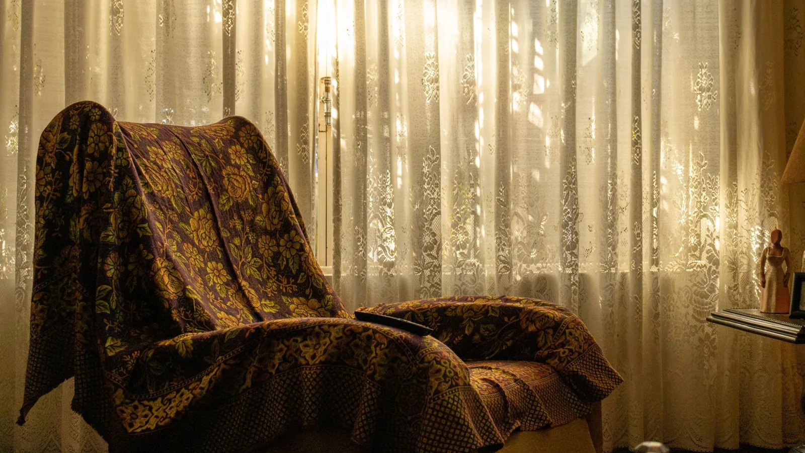 An armchair draped in a floral throw beside a window with sunlit lace curtains.