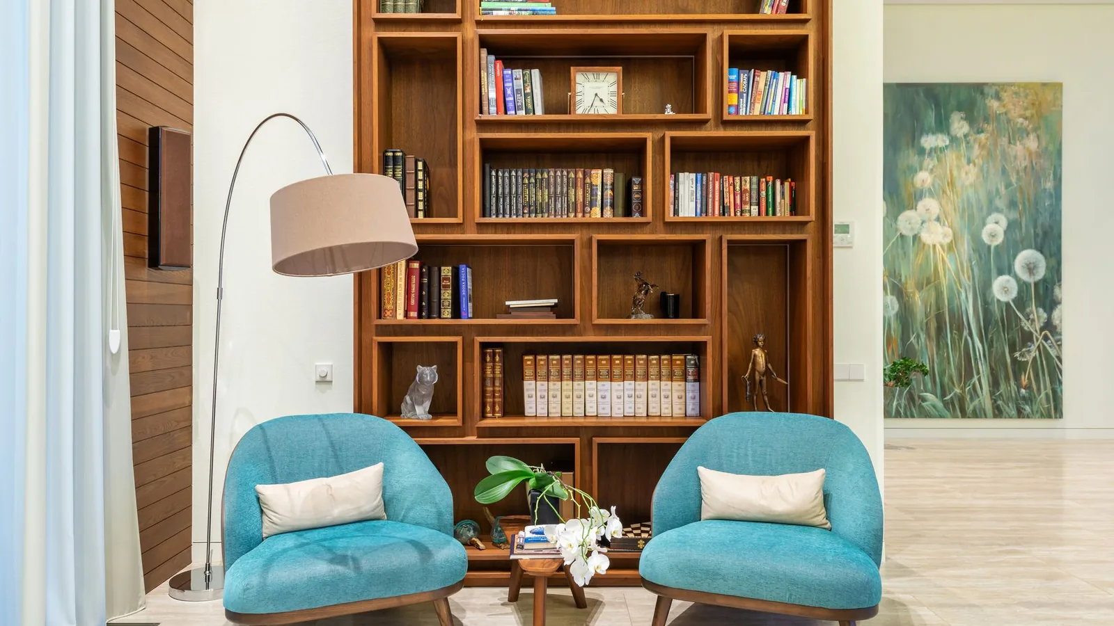 Two teal armchairs facing each other beneath a tall wooden bookcase and a floor lamp.